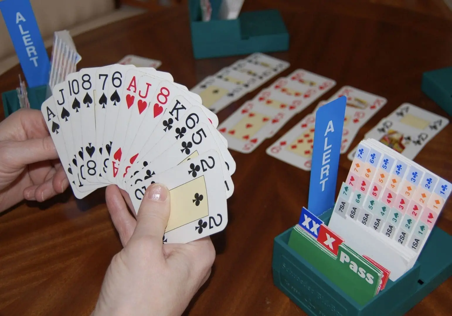 Hands holding a bridge hand of playing cards, with visible spade and heart suits, alongside an "ALERT" sign and scoring cards, set on a wooden table, highlighting the Bridge game event at Gulf Waters Beach Front RV Resort.