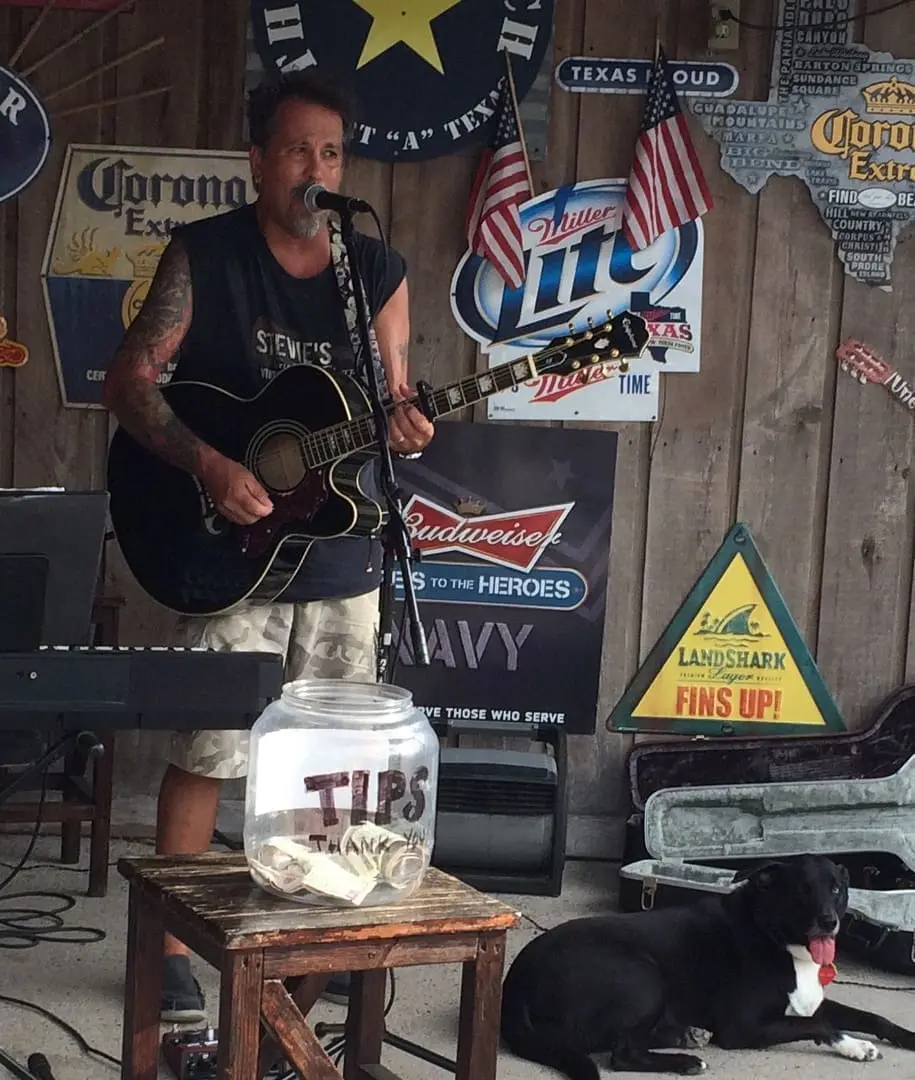 Musician Stevie Start performing live with an acoustic guitar, surrounded by bar signage, a tip jar labeled "TIPS THANK YOU," and a black dog resting nearby, reflecting the lively atmosphere of the upcoming music event at Gulf Waters Beach Front RV Resort.