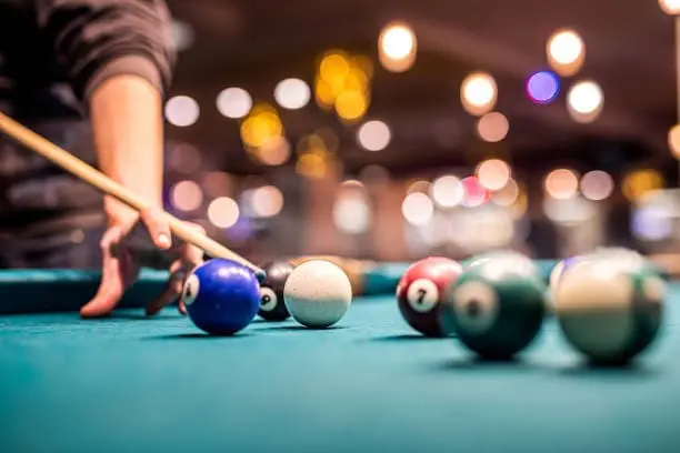 Person playing pool with cue stick, focusing on colorful billiard balls on a green felt table, in a lively indoor setting, for the Gulf Waters Pool Tournament event.