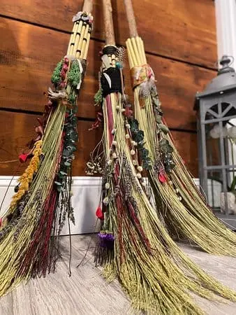 Halloween-themed decorative brooms with colorful embellishments, displayed against a wooden background, promoting the broom-making class at Gulf Waters Beach Front RV Resort.