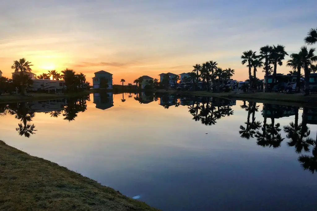 ### Key Elements

#### Main Subjects
- Sunset over a water body
- Reflection of buildings and palm trees

#### Objects
- Palm trees lining the shore
- Buildings with a reflective surface on the water

#### Notable Features
- Clear, calm water creating a mirror-like effect
- Vibrant colors in the sky transitioning from orange to blue

#### Text or Symbols
- None present

### Overall Mood or Theme
The image evokes a serene and tranquil atmosphere, capturing a peaceful evening scene with natural beauty and harmonious reflections, suggesting relaxation and calmness.