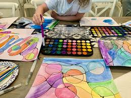 Child painting with watercolors at an art class table, surrounded by colorful artworks and a palette of vibrant paints, reflecting the creative atmosphere of the Gulf Waters Beach Front RV Resort art class event.