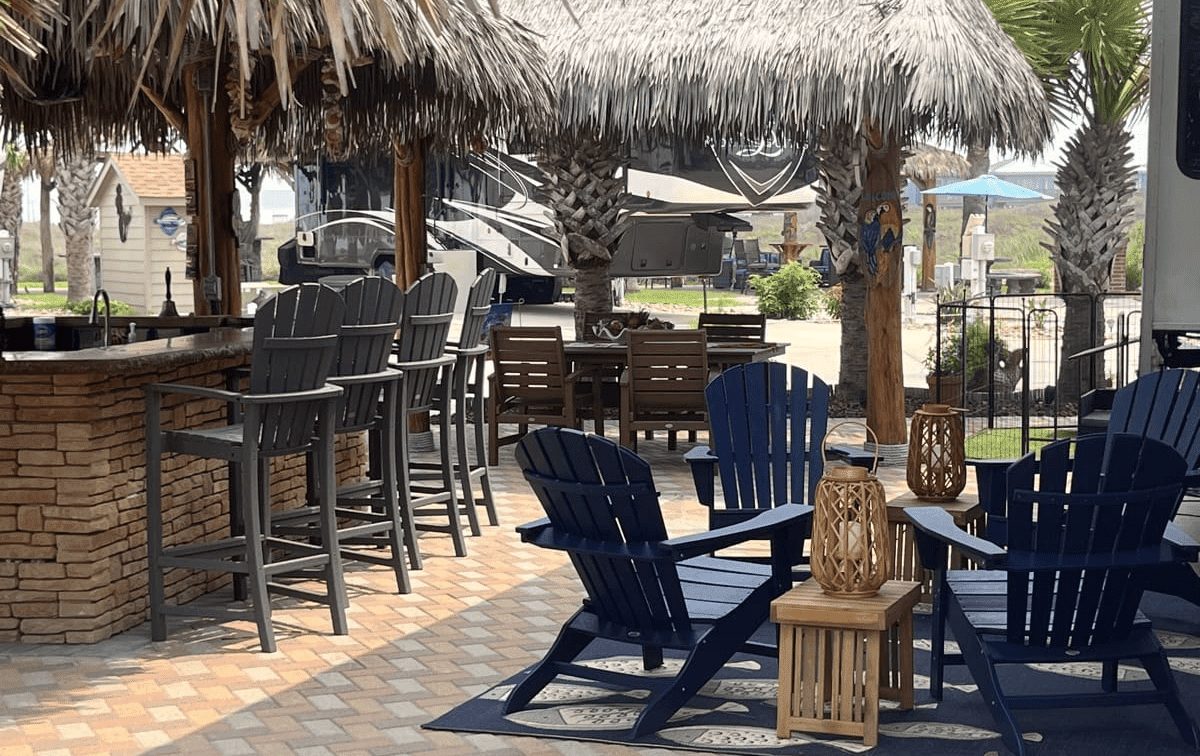 Outdoor lounge area at Gulf Waters Beach Front RV Resort featuring blue adirondack chairs, a tiki-style bar, and tropical landscaping, ideal for relaxation and socializing near the Texas Gulf Coast.