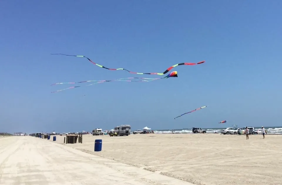 Colorful kites flying over a sandy beach with people and vehicles nearby.