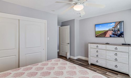 Cozy bedroom in a beach house at Gulf Waters Beach Front RV Resort, featuring a queen bed with patterned bedding, a dresser, a ceiling fan, and a wall-mounted TV displaying scenic views.