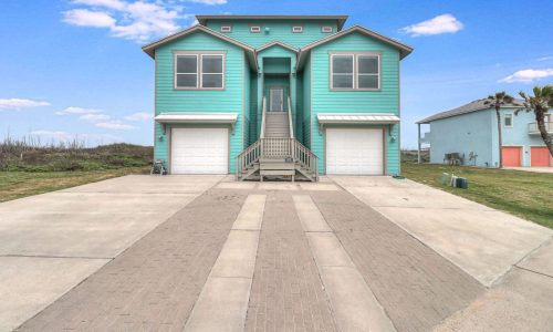 Beach house rental at Gulf Waters RV Resort, featuring a turquoise exterior, large windows, and a welcoming entrance with stairs, surrounded by green grass and blue sky.