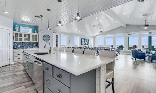 Modern beach house interior with open layout, featuring a large kitchen island, coastal decor, and panoramic windows overlooking Mustang Island's sandy shores at Gulf Waters RV Resort.