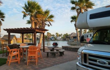 Luxury RV site at Gulf Waters Beach Front RV Resort, featuring palm trees, outdoor seating area with a pergola, and scenic pond view, emphasizing coastal charm and relaxation amenities.