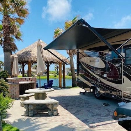 Beachfront RV site with palm trees, picnic table, and a shaded area featuring a thatched roof, showcasing Gulf Waters Beach Front RV Resort amenities.
