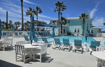 Heated pool area at Gulf Waters Beach Front RV Resort with white lounge chairs, blue umbrellas, and tropical palm trees, showcasing resort-style amenities and coastal charm.