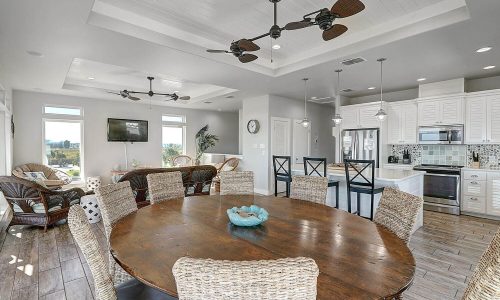 Spacious dining area with a large wooden table and wicker chairs, featuring modern kitchen appliances, ceiling fans, and large windows offering views, showcasing the inviting interior of a beach house at Gulf Waters Beach Front RV Resort.