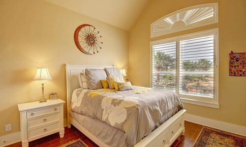 Cozy beach house bedroom with a queen bed, decorative pillows, bedside lamps, and large windows showcasing palm trees, reflecting the modern comforts of Gulf Waters Beach Front RV Resort accommodations.