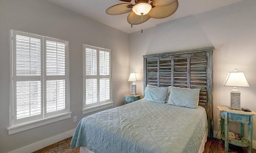 Cozy beach house bedroom featuring a queen-sized bed with light blue bedding, wooden headboard, and decorative lamps, complemented by natural light from two windows, ideal for a relaxing stay at Gulf Waters Beach Front RV Resort.