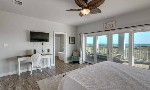 Beach house interior featuring a cozy bedroom with a large bed, a flat-screen TV, a desk, and sliding glass doors leading to a deck with ocean views, emphasizing modern comforts and beach getaway ambiance.