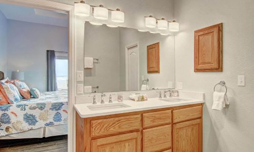 Bathroom interior with double sink, wooden cabinetry, and a mirror, featuring coastal decor and a view into a bedroom with a bed adorned in colorful bedding, reflecting the beach house rental experience at Gulf Waters Beach Front RV Resort.