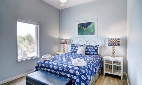 Cozy bedroom in a beach house rental at Gulf Waters RV Resort, featuring blue and white decor, a comfortable bed with seashell-patterned bedding, bedside tables with lamps, and a window with a view of palm trees, perfect for a coastal getaway.