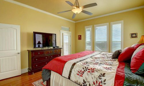 Cozy bedroom in a Gulf Waters Beach Front RV Resort beach house, featuring a king-sized bed with floral bedding, a dresser, a flat-screen TV, and large windows offering natural light and views.