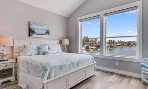 Cozy beach house interior featuring a queen bed with light blue bedding, large windows offering views of the water and palm trees, and a serene coastal decor, ideal for vacation rentals at Gulf Waters RV Resort.