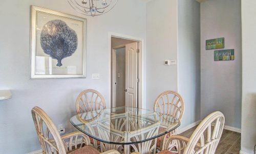 Dining area in a beach house rental featuring a glass table surrounded by rattan chairs, coastal-themed artwork on the walls, and a bright, airy atmosphere, emphasizing modern comforts for vacationers at Gulf Waters Beach Front RV Resort.