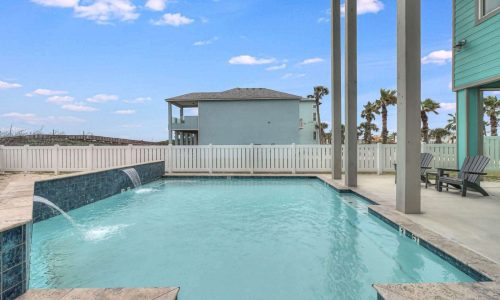 Beach house rental pool area with clear blue water, palm trees, and adjacent house in Gulf Waters RV Resort, perfect for a coastal escape on Mustang Island.