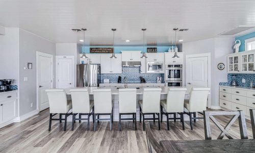 Modern kitchen interior of a beach house rental at Gulf Waters RV Resort, featuring white cabinetry, stainless steel appliances, and a large dining table with white chairs, emphasizing comfort and style for vacationers.