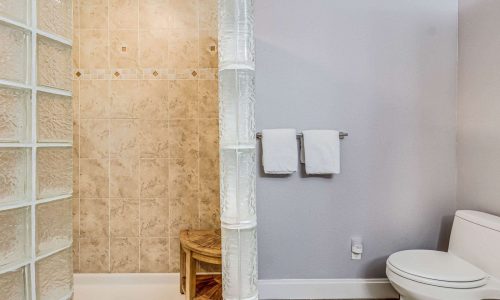 Bathroom interior featuring a glass block shower, beige tiled walls, wooden stool, and white towels, highlighting modern comforts in a beach house at Gulf Waters Beach Front RV Resort.