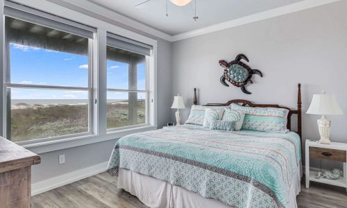 Cozy beach house bedroom with large windows showcasing coastal views, featuring a king-size bed with patterned bedding, decorative turtle wall art, and bedside tables, emphasizing a relaxing atmosphere for vacationers at Gulf Waters Beach Front RV Resort.