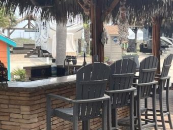 Bar seating area with gray Adirondack chairs under a thatched roof, overlooking a beachfront RV resort setting at Gulf Waters Beach Front RV Resort.