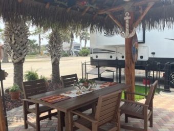 Outdoor dining area with wooden table and chairs under a thatched roof, surrounded by palm trees and RV parking, highlighting the resort's amenities at Gulf Waters Beach Front RV Resort.