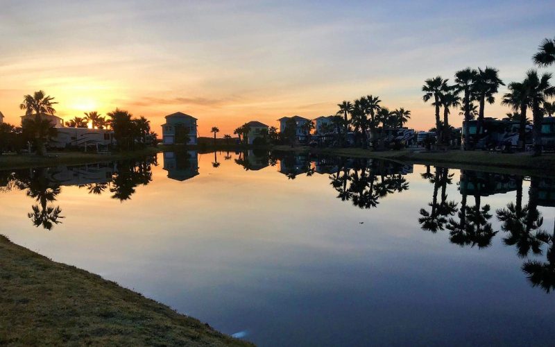 ### Key Elements

#### Main Subjects
- Sunset over a water body
- Reflection of buildings and palm trees

#### Objects
- Palm trees lining the shore
- Buildings with a reflective surface on the water

#### Notable Features
- Clear, calm water creating a mirror-like effect
- Vibrant colors in the sky transitioning from orange to blue

#### Text or Symbols
- None present

### Overall Mood or Theme
The image evokes a serene and tranquil atmosphere, capturing a peaceful evening scene with natural beauty and harmonious reflections, suggesting relaxation and calmness.