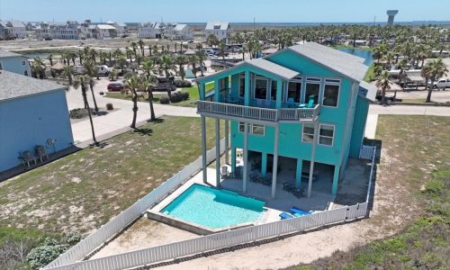 Aerial view of a vibrant turquoise beach house with a private pool, situated near Mustang Island, surrounded by palm trees and vacation homes at Gulf Waters Beach Front RV Resort.
