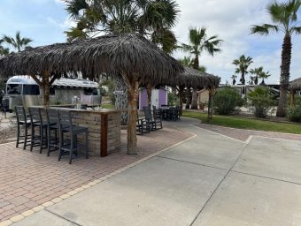 Beachfront RV resort amenities with thatched-roof cabanas, outdoor seating, palm trees, and paved pathways at Gulf Waters Beach Front RV Resort in Port Aransas, Texas.