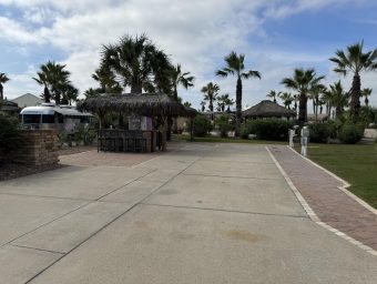 Beachfront RV resort entrance featuring palm trees, a thatched-roof cabana, and paved pathways, highlighting Gulf Waters Beach Front RV Resort amenities.