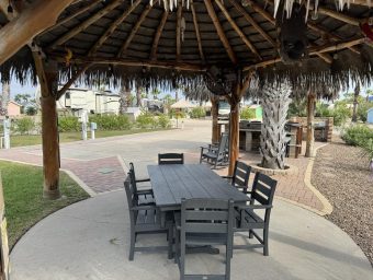 Outdoor seating area under a thatched roof gazebo at Gulf Waters Beach Front RV Resort, featuring a large table and chairs, surrounded by landscaped grounds and RV lots.