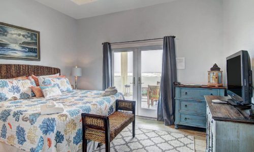 Cozy beach house interior featuring a king-sized bed with ocean-themed bedding, a dresser, and a TV, with patio doors leading to an outdoor seating area, ideal for a relaxing stay at Gulf Waters Beach Front RV Resort.