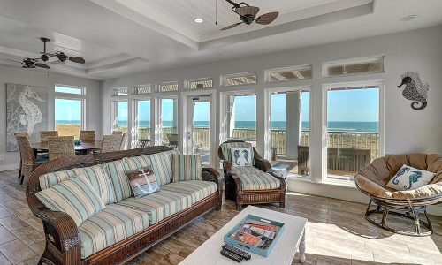 Bright and airy beach house interior with large windows showcasing ocean views, featuring a striped sofa, wicker chairs, and a dining area, embodying modern coastal decor for a relaxing getaway at Gulf Waters Beach Front RV Resort.