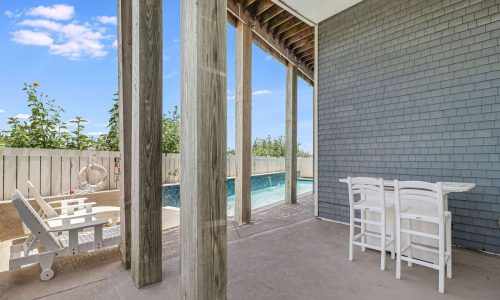 Private outdoor patio area of a beach house at Gulf Waters Beach Front RV Resort, featuring white furniture, wooden beams, and a view of a swimming pool, ideal for relaxing during a beach getaway.