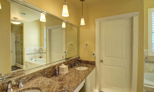 Modern bathroom with dual sinks, granite countertop, and large mirror, featuring warm lighting and a door leading to a shower area, reflecting the comfort and style of Gulf Waters Beach Front RV Resort accommodations.