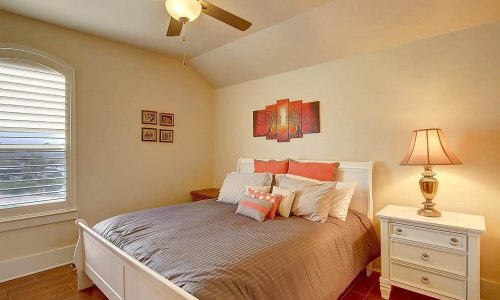 Cozy bedroom in a Gulf Waters beach house featuring a queen-sized bed with coral and white bedding, decorative pillows, a bedside lamp, and wall art, ideal for a relaxing beach getaway.
