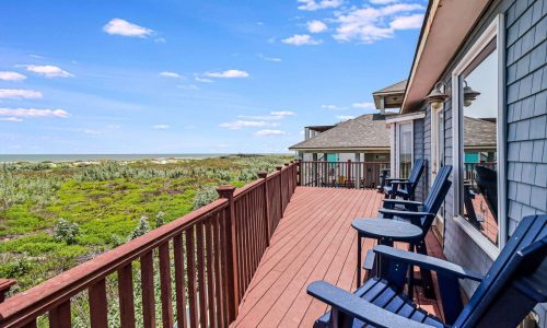 Beach house deck overlooking lush greenery and Mustang Island's sandy shores at Gulf Waters Beach Front RV Resort, featuring blue adirondack chairs for relaxation.