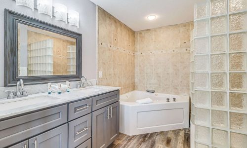 Modern bathroom featuring a double vanity with gray cabinetry, stylish fixtures, a relaxing jacuzzi tub, and glass block shower wall, reflecting the upscale accommodations at Gulf Waters Beach Front RV Resort.