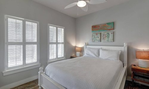 Cozy bedroom in Gulf Waters Beach Front RV Resort beach house featuring a queen bed, natural light from white-shuttered windows, and decorative artwork, ideal for relaxing beach getaways.