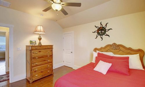 Interior of a bedroom with a wooden dresser, ceiling fan, and a bed with red bedding.