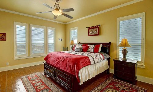 Cozy bedroom in a beach house at Gulf Waters Beach Front RV Resort, featuring a king-sized bed with red and white bedding, two bedside lamps, large windows with shutters, and a decorative area rug.