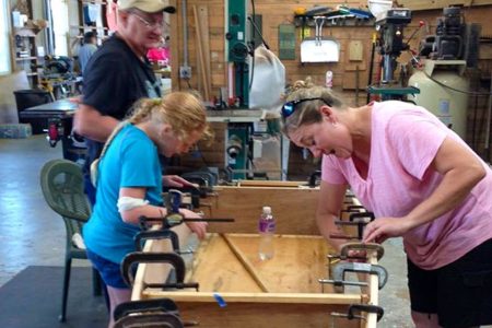 Family working together on a woodworking project in a workshop, featuring a girl and a woman using clamps on a wooden piece, with a man in the background.
