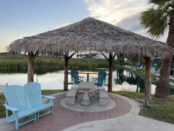 Thatched-roof gazebo with picnic table and blue Adirondack chairs by a serene waterway at Gulf Waters Beach Front RV Resort, showcasing outdoor amenities for lot owners.