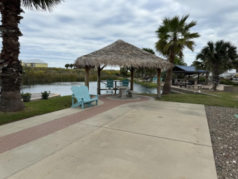 Tropical outdoor seating area with a thatched-roof gazebo, blue adirondack chairs, and a scenic pond, highlighting amenities at Gulf Waters Beach Front RV Resort.