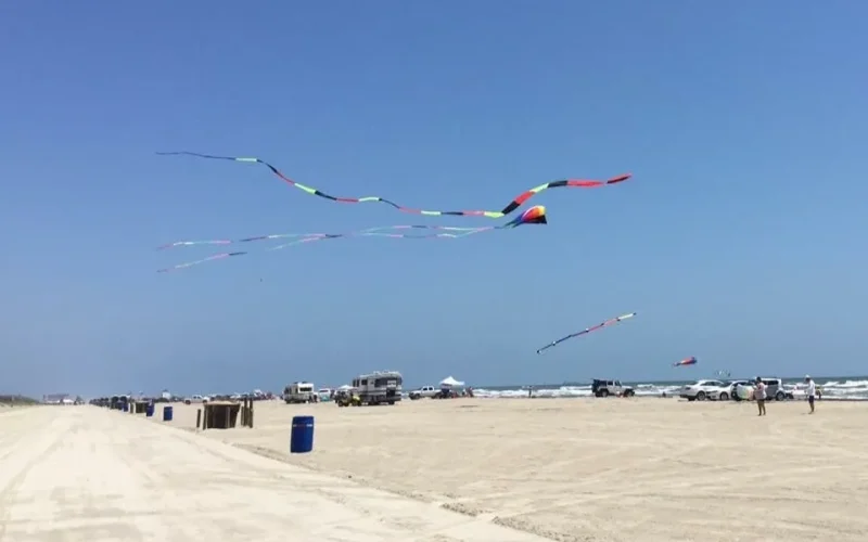 Colorful kites flying over a sandy beach with people and vehicles nearby.