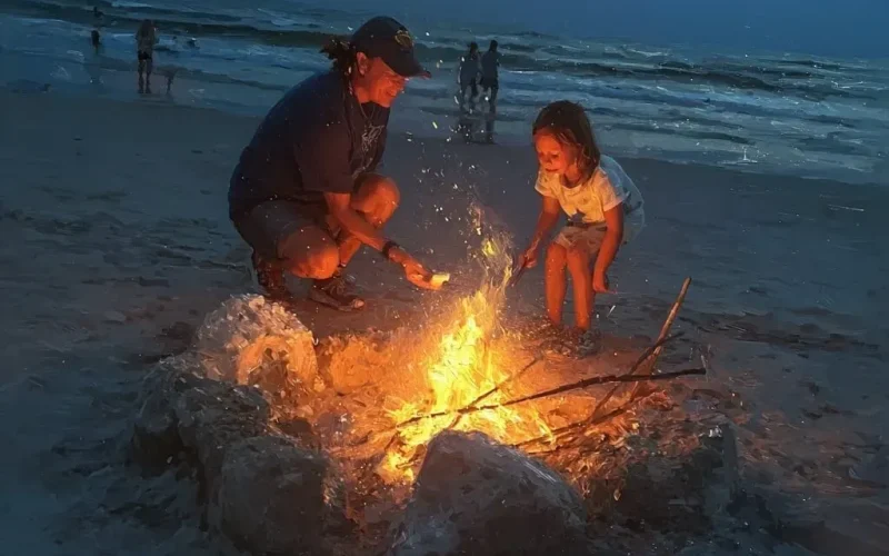 Child and adult gathering around a beach bonfire at sunset.