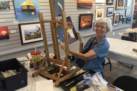 Woman painting on an easel in the Port Aransas Art Center, surrounded by art supplies and framed artworks on display.